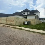 A wooden fence constructed from vertical planks, showcasing a natural wood finish, sturdy design, and surrounding cement driveway and yard filled with green grass.