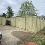 A wooden fence constructed from vertical planks, showcasing a natural wood finish, sturdy design, and surrounding yard filled with green grass and a cement walkway.