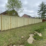 A wooden fence constructed from vertical planks, showcasing a natural wood finish, sturdy design, and surrounding yard filled with green grass.