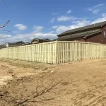 A wooden fence constructed from vertical planks, showcasing a natural wood finish, sturdy design, and surrounding yard filled with dirt.