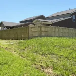A wooden fence constructed from vertical planks, showcasing a natural wood finish, sturdy design, and surrounding yard filled with green grass.
