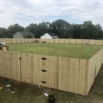 A fence constructed from iron, showcasing a natural wood finish, sturdy design, and two single-story homes with a surrounding yard filled with green grass.