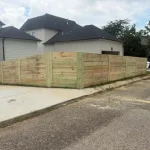 A wooden fence constructed from vertical planks, showcasing a natural wood finish, sturdy design, and surrounding cement driveway and yard filled with green grass.