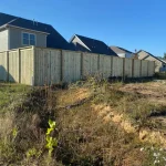 A wooden fence constructed from vertical planks, showcasing a natural wood finish, sturdy design, and surrounding yard filled with tall unkempt grass.