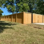 A wooden fence constructed from vertical planks, showcasing a natural wood finish, sturdy design, and surrounding yard filled with unkempt grass.