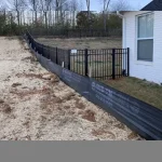 A fence constructed from iron, showcasing a natural wood finish, sturdy design, and surrounding yard filled with dirt and a single story home.