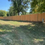 A wooden fence constructed from vertical planks, showcasing a natural wood finish, sturdy design, and surrounding yard filled with green grass.
