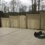 A wooden fence constructed from vertical planks, showcasing a natural wood finish, sturdy design, and surrounding cement driveway and yard filled with green grass.