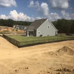 A fence constructed from iron, showcasing a natural wood finish, sturdy design, and surrounding a farmhouse and yard filled with green grass.