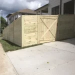 A wooden fence and gate constructed from vertical planks, showcasing a natural wood finish, sturdy design, and surrounding yard filled with green grass and a cement driveway.