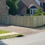 A wooden fence constructed from vertical planks, showcasing a natural wood finish, sturdy design, and surrounding cement driveway and yard filled with green grass.