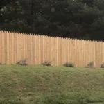 A wooden fence constructed from vertical planks, showcasing a natural wood finish, sturdy design, and surrounding yard filled with green grass.