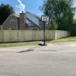 A wooden fence constructed from vertical planks, showcasing a natural wood finish, sturdy design, and surrounding cement driveway and yard filled with green grass. A basketball hoop is in the street.
