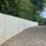 A wooden fence constructed from vertical planks, showcasing a natural wood finish, sturdy design, and surrounding yard filled with dirt.