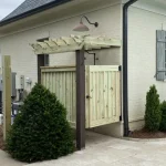 A wooden patio constructed from vertical planks, showcasing a natural wood finish, sturdy design, and surrounding cement driveway and yard filled with green bushes and cement walkways.