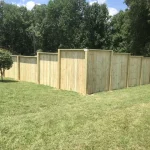 A wooden fence constructed from vertical planks, showcasing a natural wood finish, sturdy design, and surrounding yard filled with green grass.