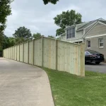 A wooden fence constructed from vertical planks, showcasing a natural wood finish, sturdy design, and surrounding yard filled with green grass and a cement walkway.