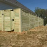 A wooden fence constructed from vertical planks, showcasing a natural wood finish, sturdy design, and surrounding yard filled with dirt.